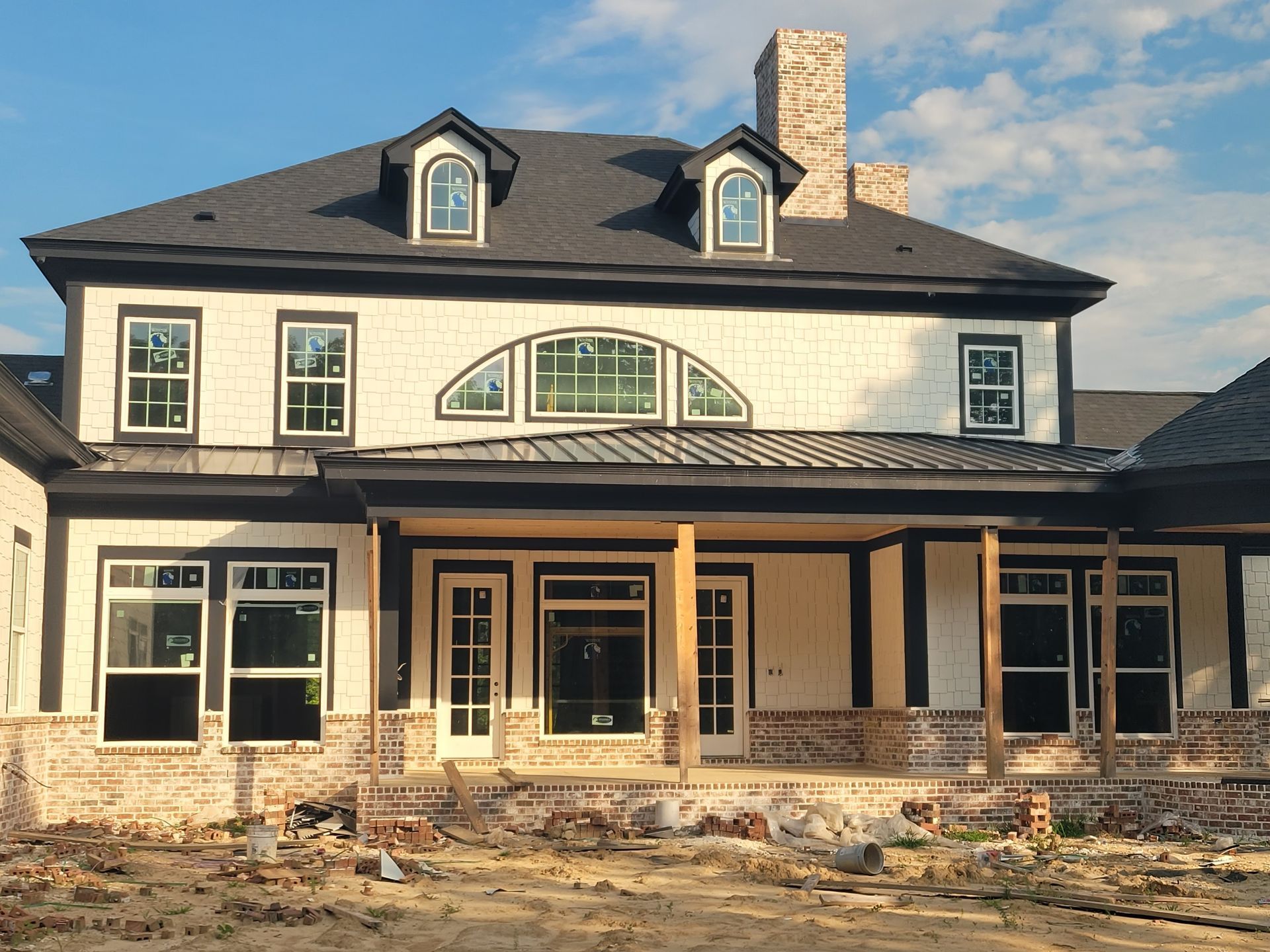 Two-story house under construction, white siding, black trim, brick base, arched window, blue sky.