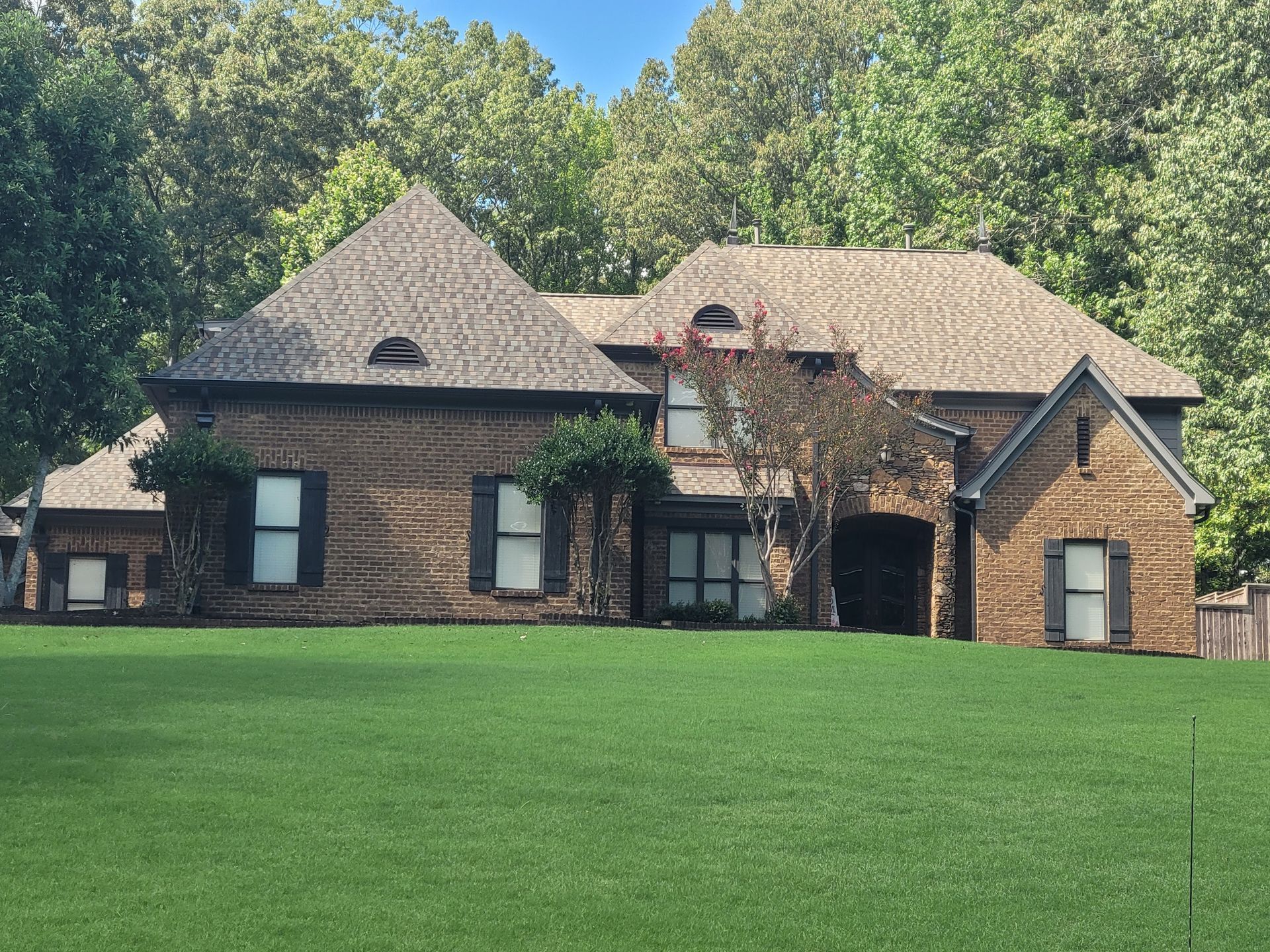 Brick house with a green lawn in front of trees. Dark shutters and tan roof.