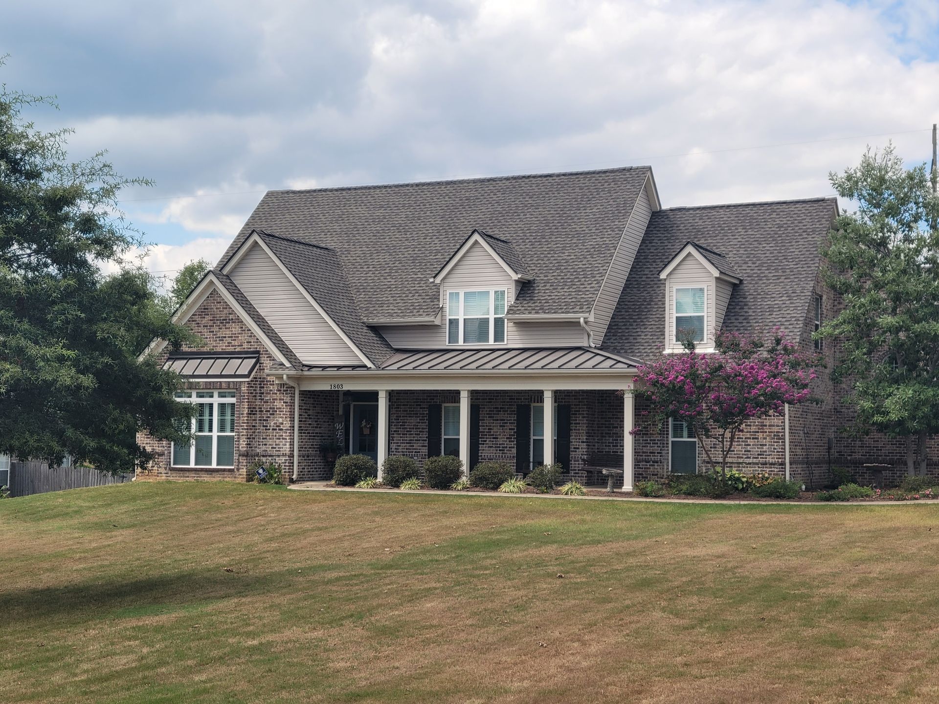 Two-story brick home with a gray roof and a covered porch on a grassy lawn under a cloudy sky.