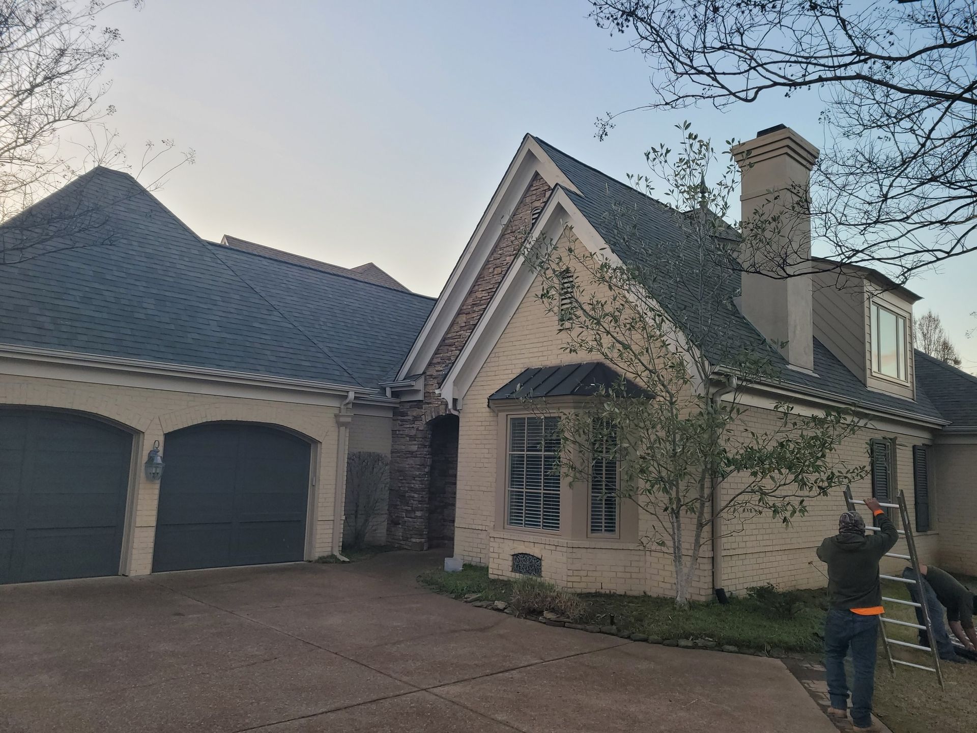 Tan house with dark green roof, gray garage doors, and brick accents. Two people work on a ladder.