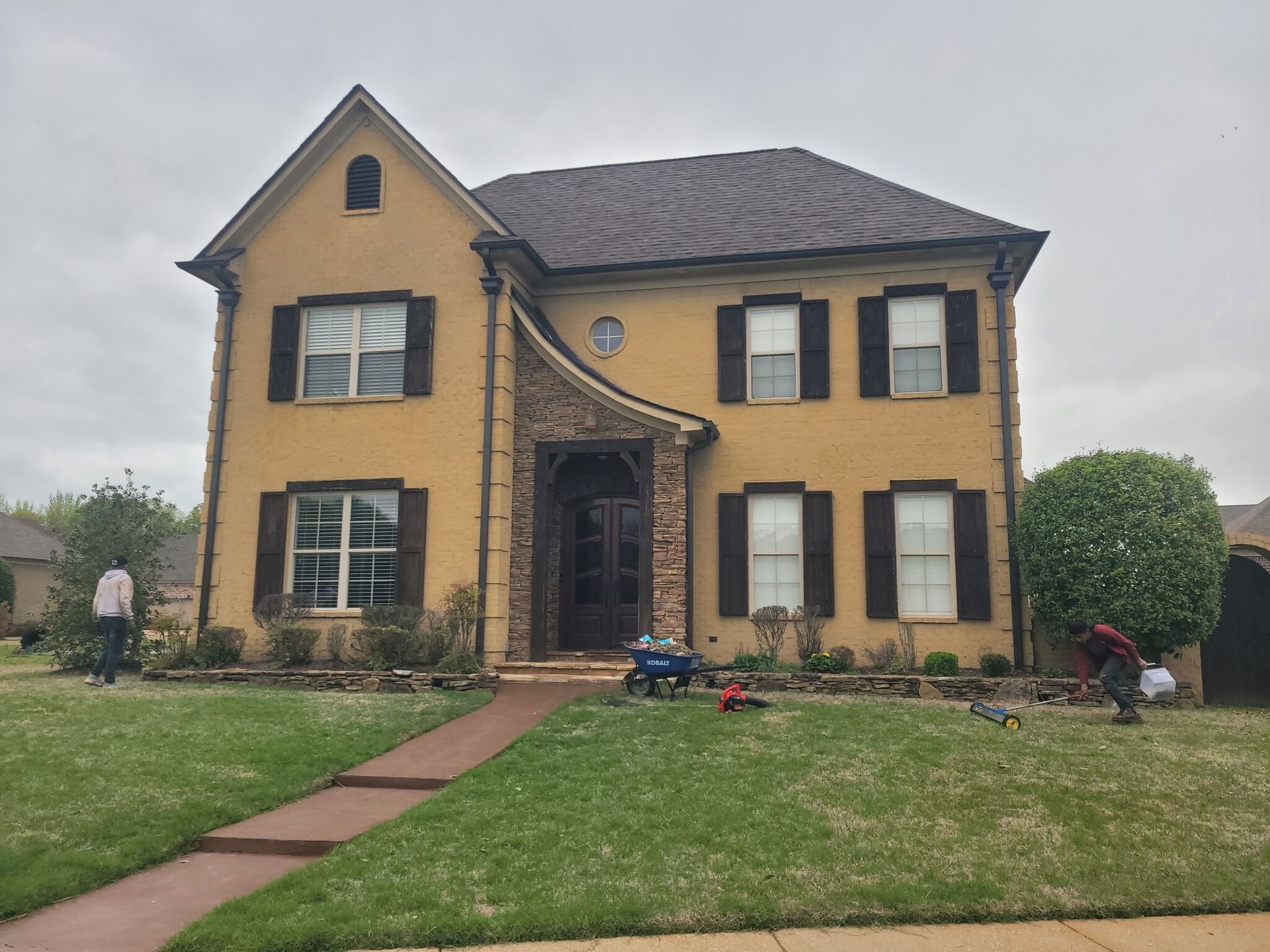 Yellow two-story house with dark shutters and brown roof. People are working on the lawn.