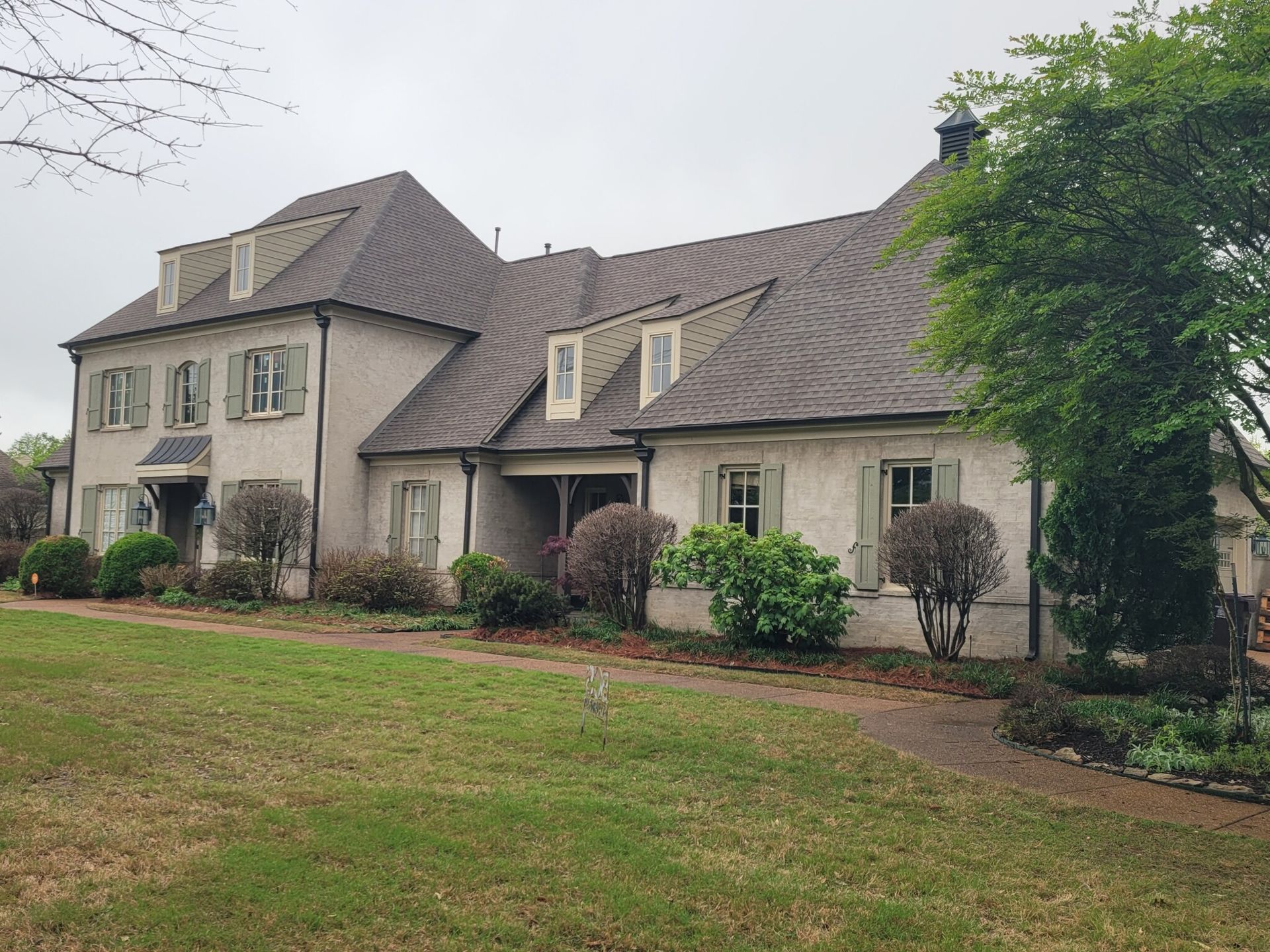 A two-story stucco house with a brown roof, green shutters, and dormant landscaping on a cloudy day.