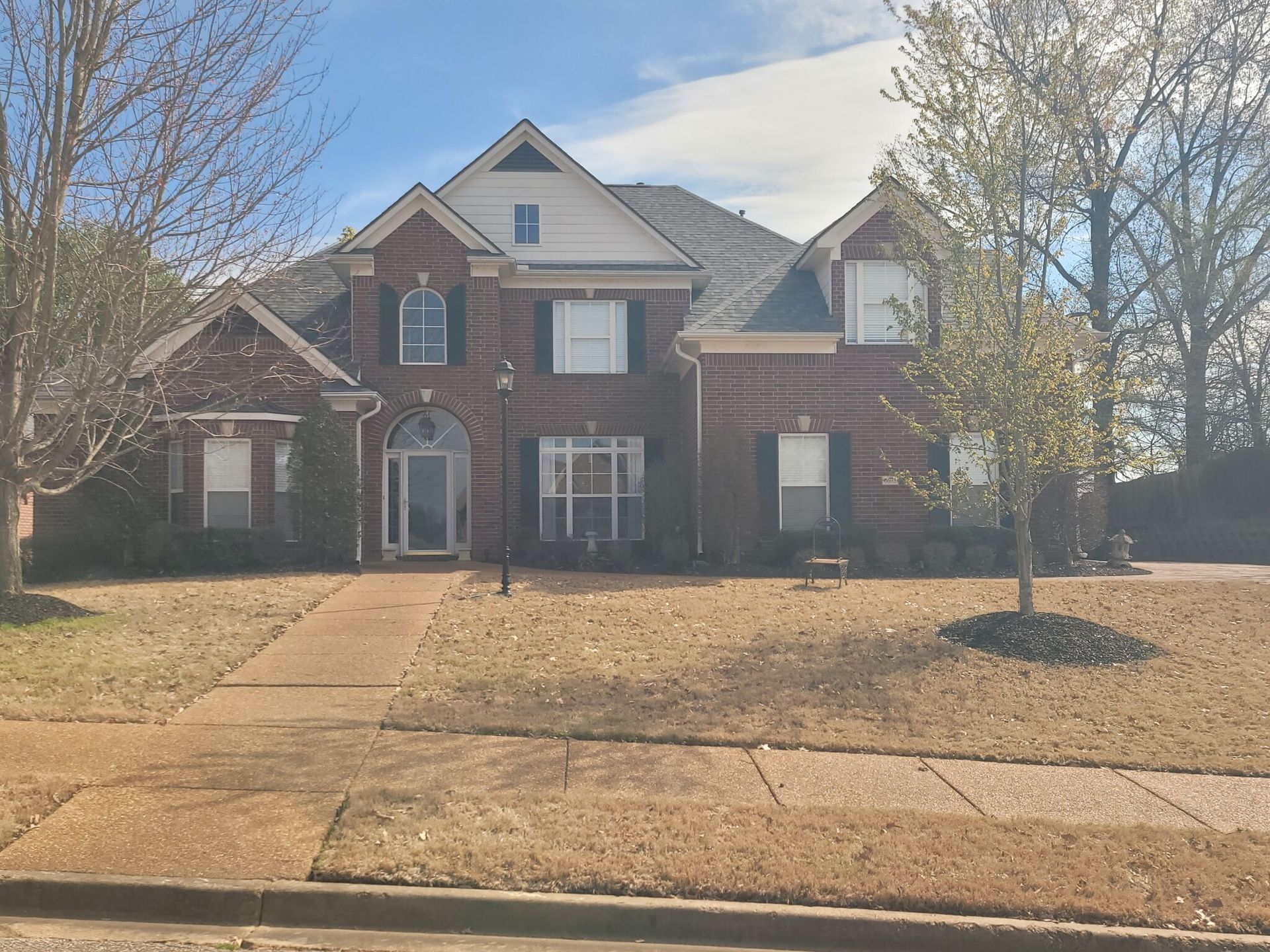 Red brick house with black shutters, dry brown grass in the yard, and leafless trees.