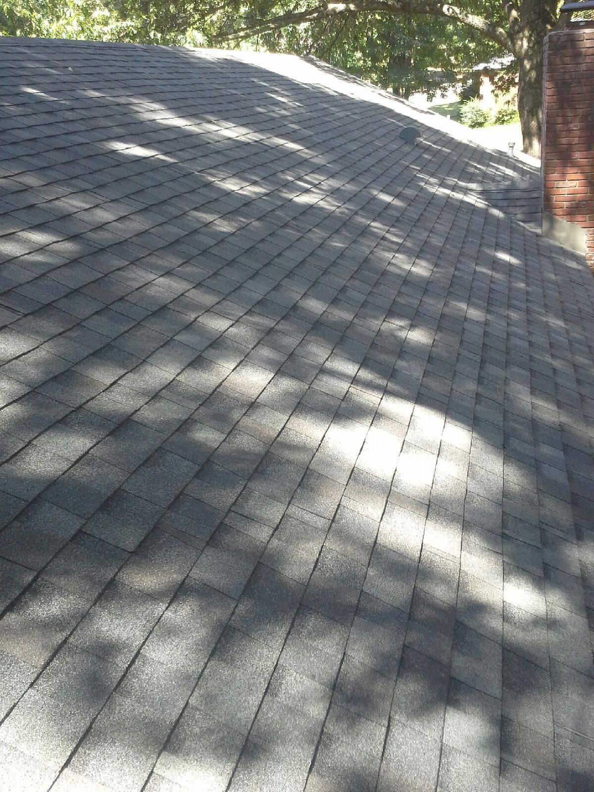 A dark gray shingle roof with shadows from trees, next to a brick chimney.
