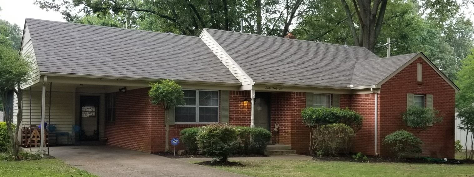 A red brick house with a gray roof, carport, and green lawn and shrubs.