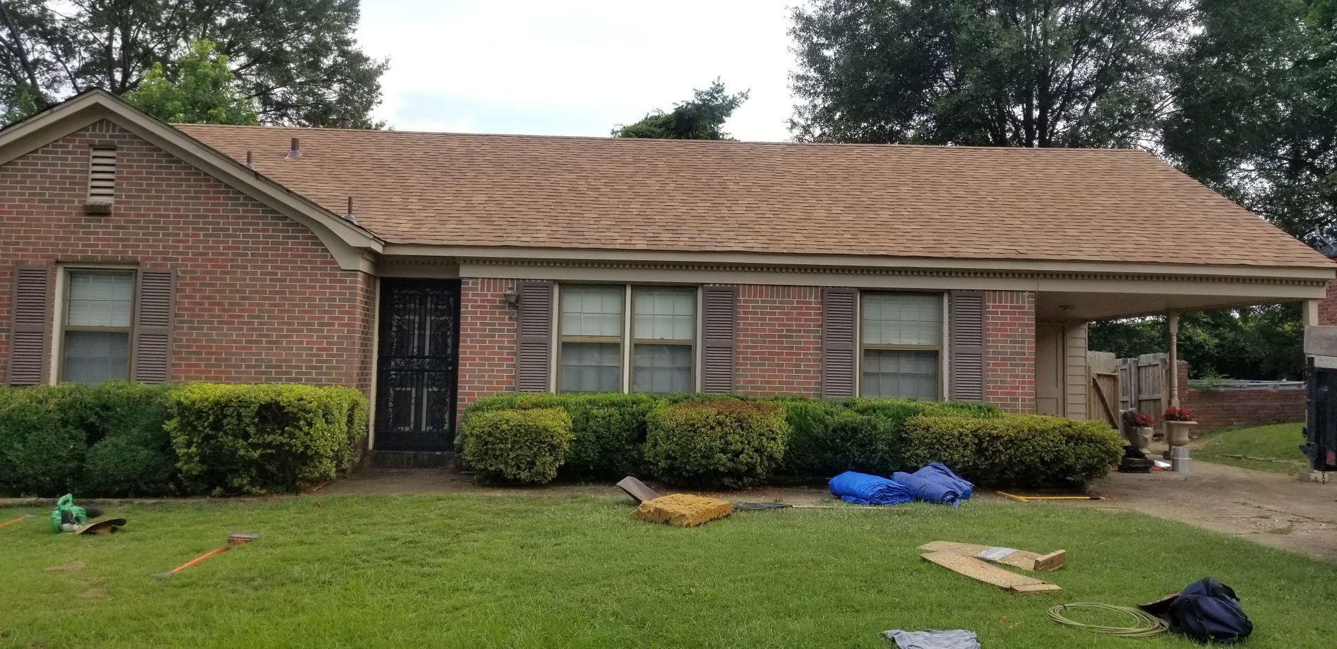 Brick house with a partially torn-off roof; yard with bushes, blue tarp, and debris.