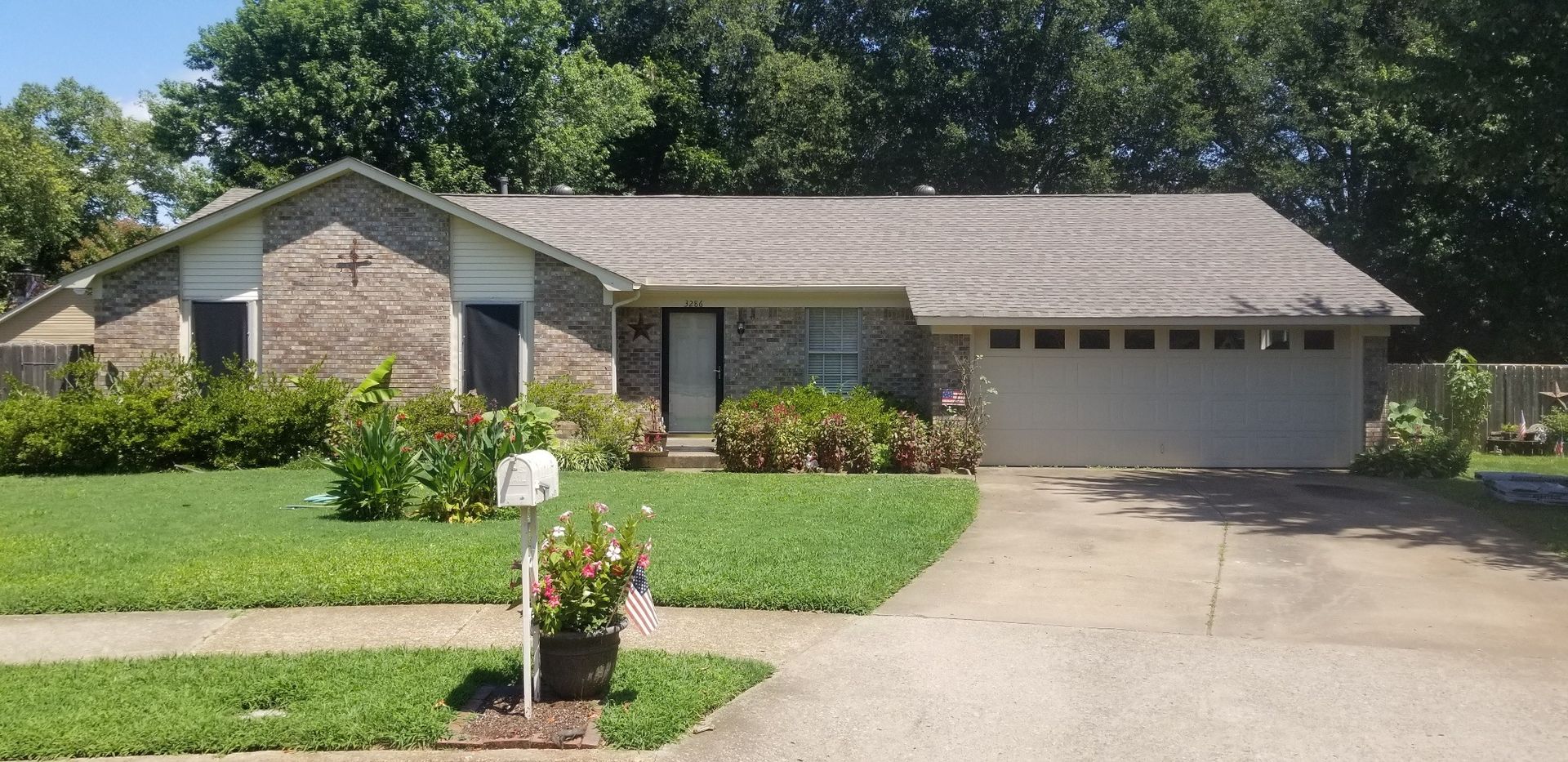 A single-story brick house with a gray roof, lawn, and driveway. Trees are in the background.
