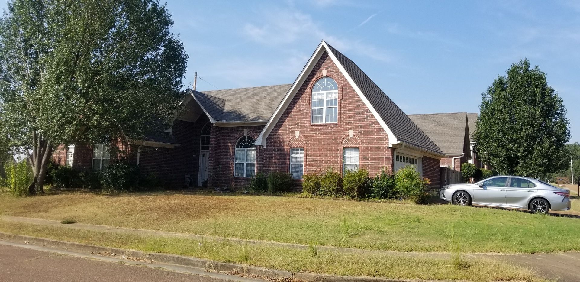 A brick house with a large tree in front and a silver car parked on the lawn.