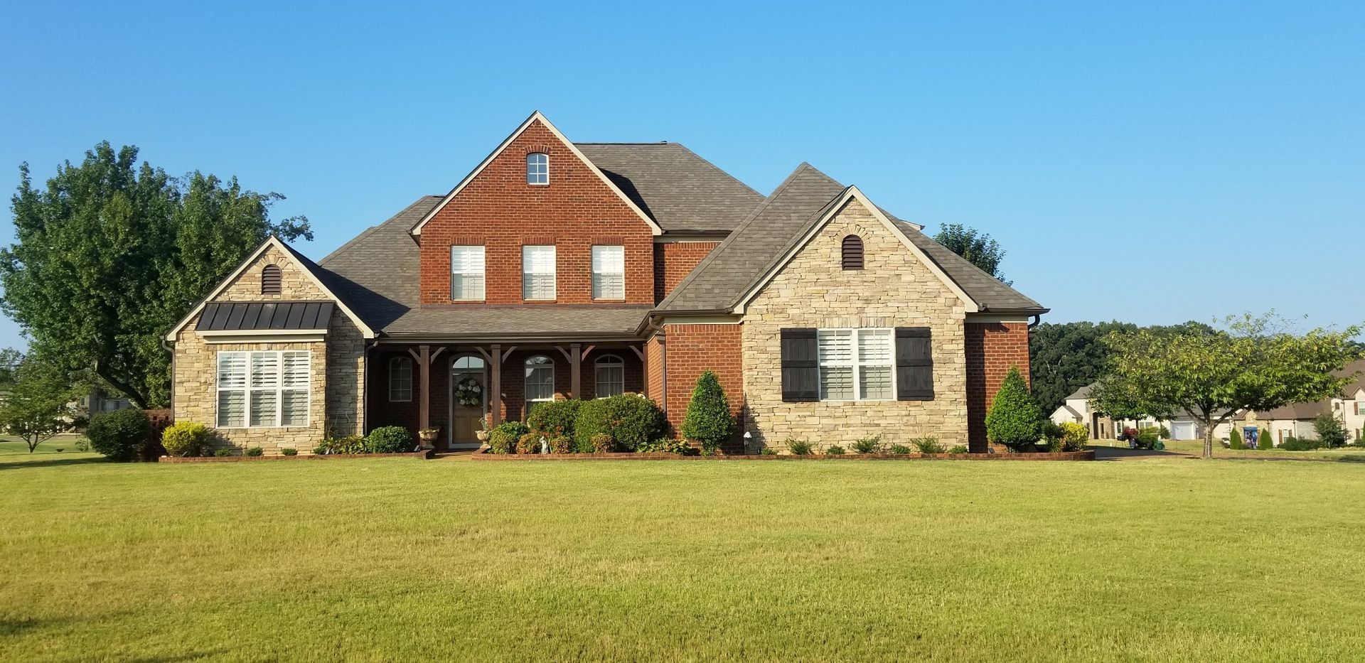 Large two-story house with a brick and wood facade, set on a large lawn under a blue sky.