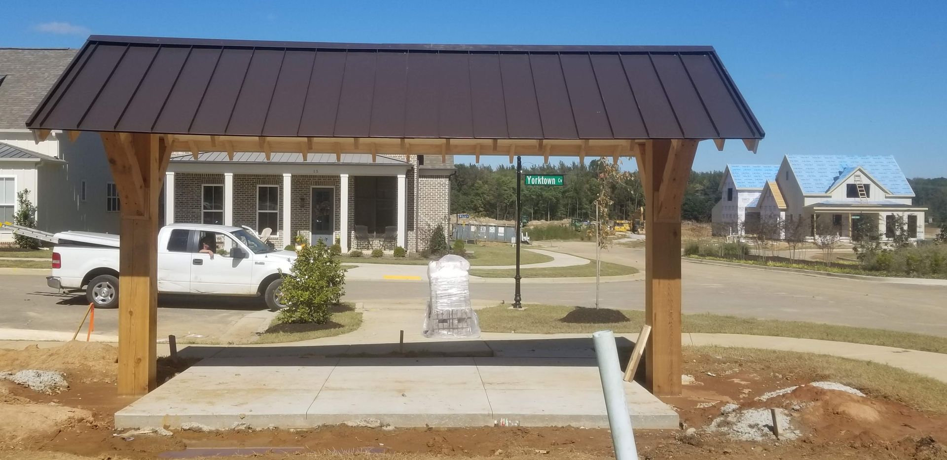 Wooden shelter with brown metal roof; white truck; new homes on a sunny day.