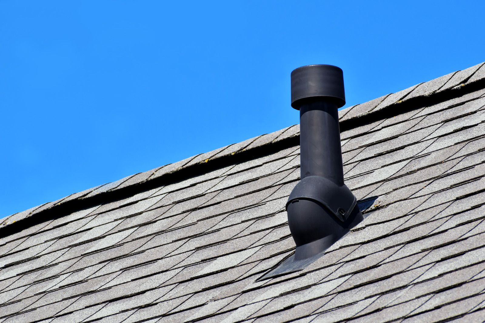 Black vent pipe on a shingle roof against a clear blue sky.