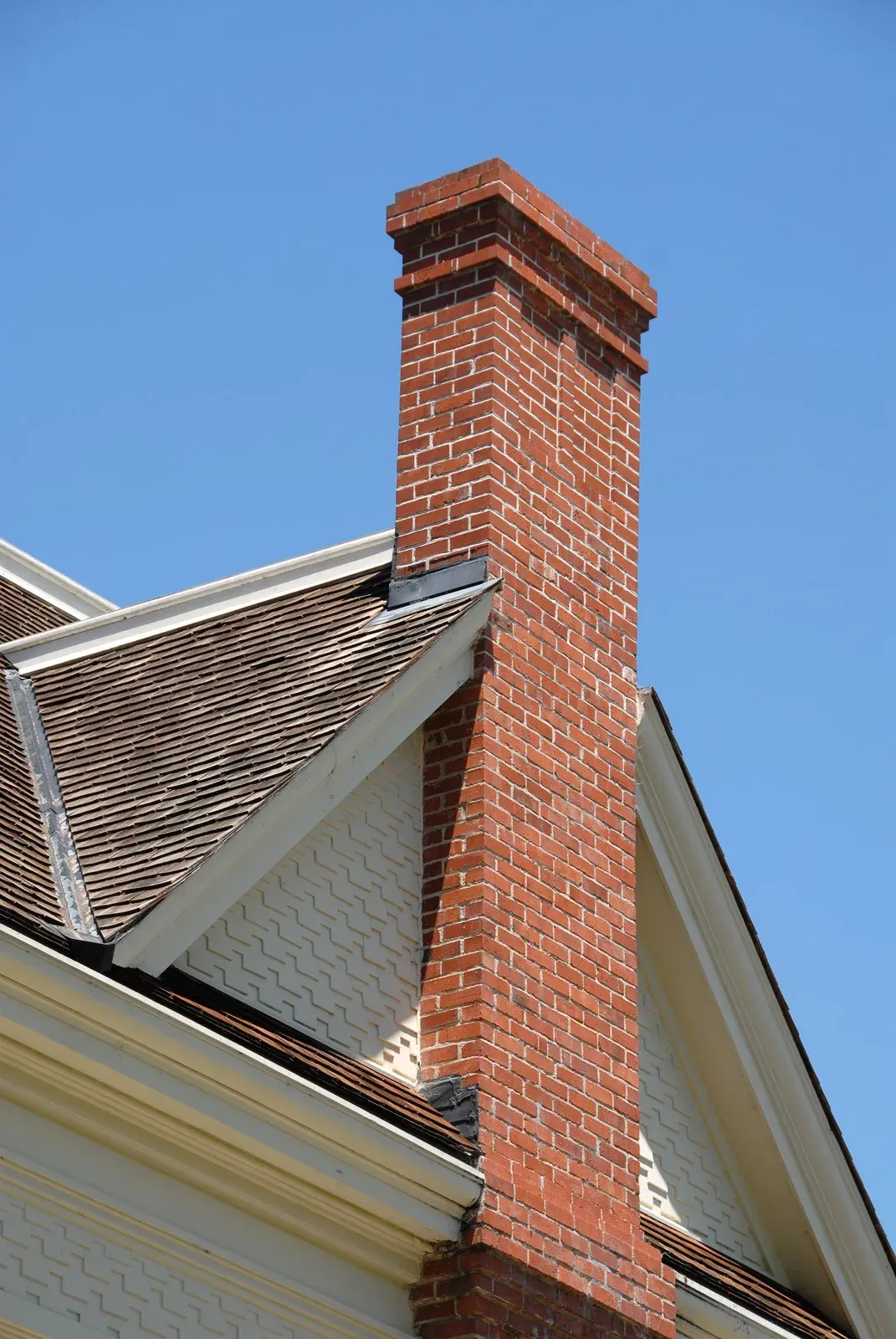 Brick chimney against a blue sky, rising from a gabled roof with brown shingles.