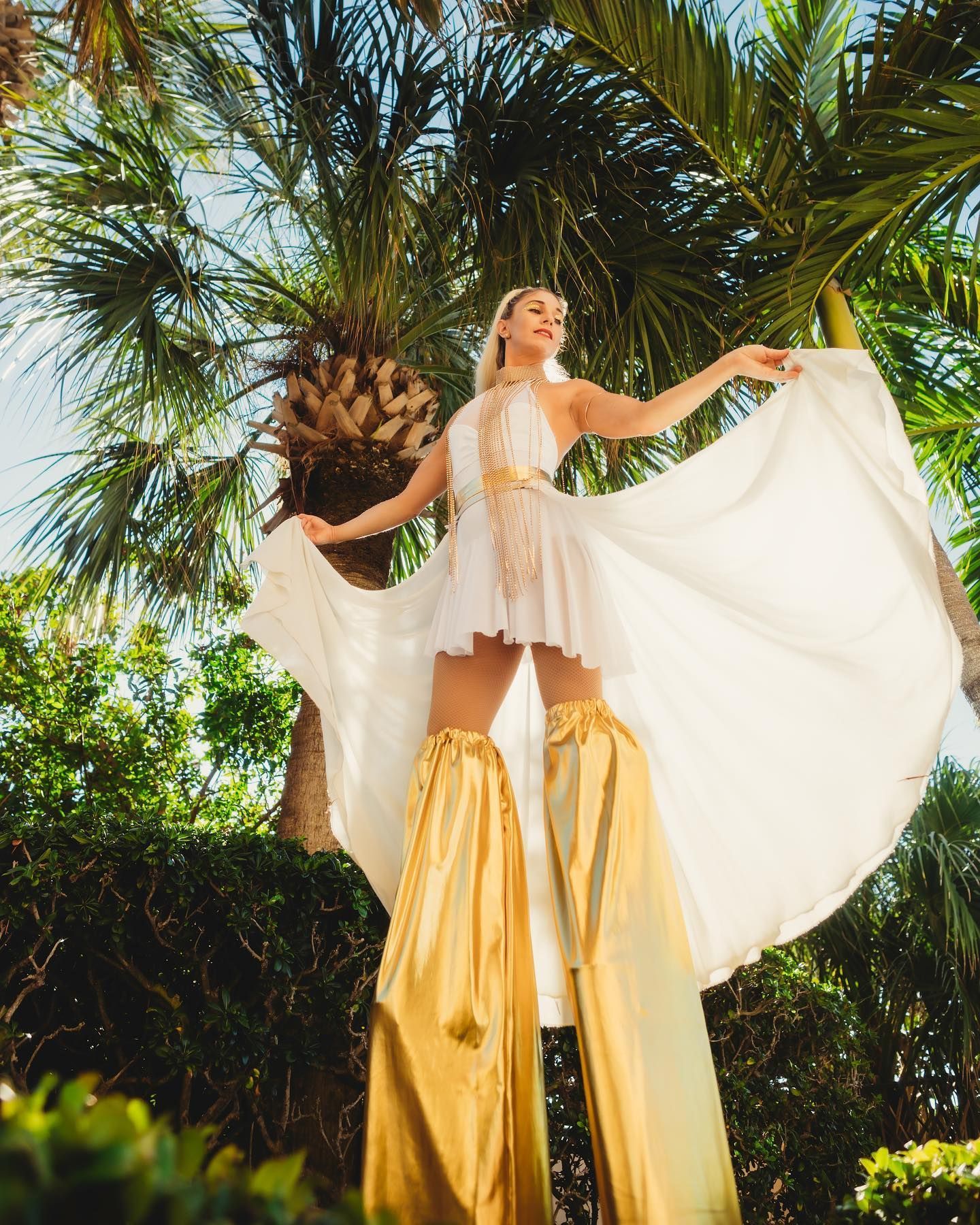 A woman in a white dress and gold stilts is standing in front of a palm tree.