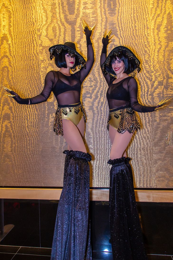 Two women are standing on stilts in front of a gold wall.