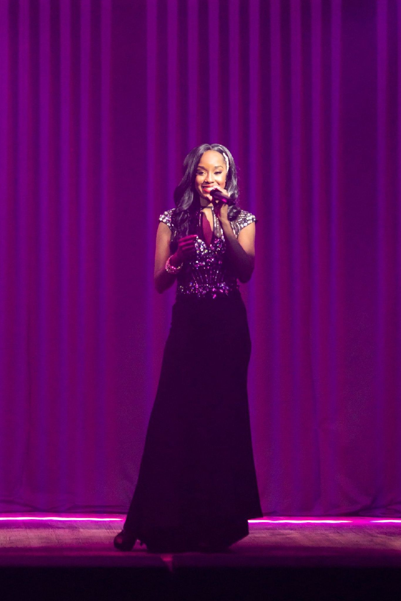 A woman in a long black dress is talking into a microphone on a stage.
