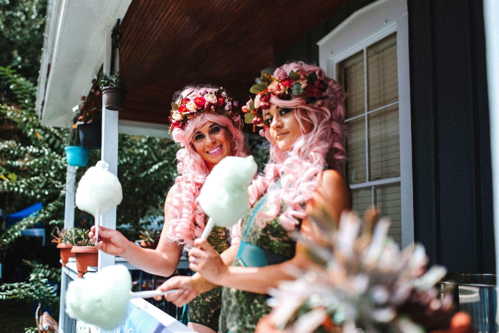 Two women in pink wigs have cotton candy on a porch.