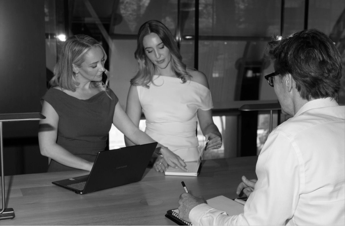 Three professionals in an office collaborate around a desk while reviewing documents on a laptop and paper.