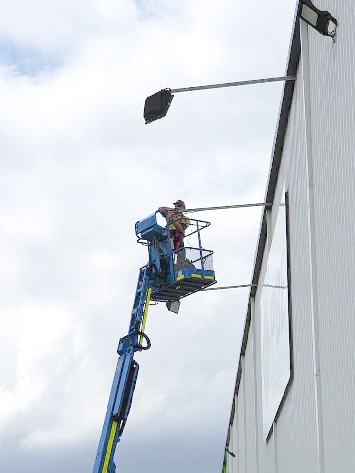 Person on a blue lift working on a light fixture attached to a white building under a cloudy sky — Firefly Connections In Teralba, NSW