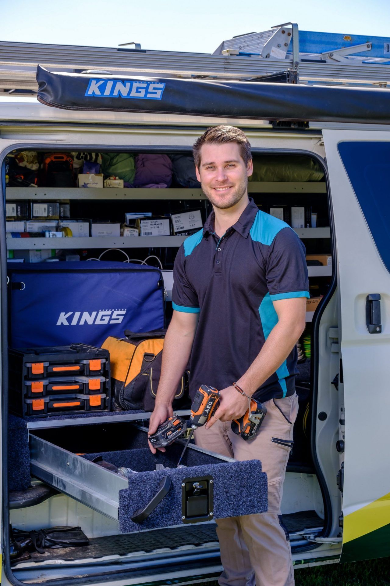 Man in work attire smiling, holding a drill, standing in front of a utility van with tools and supplies — Firefly Connections In Mayfield, NSW