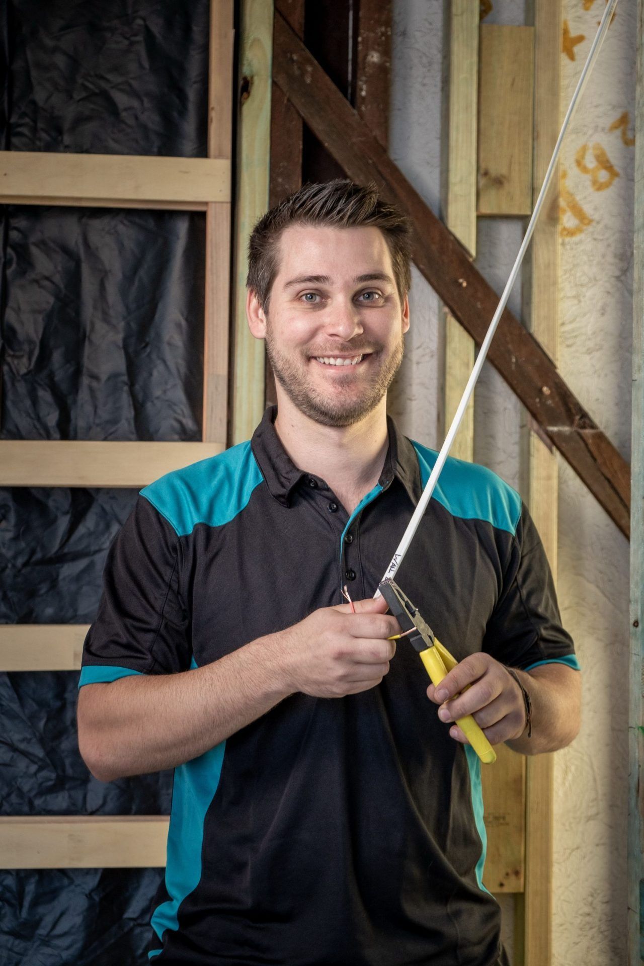 Smiling man in teal and black shirt holding pliers against a construction background — Firefly Connections In Teralba, NSW