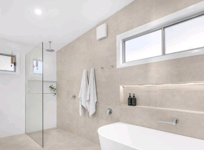 Modern bathroom with neutral beige tiles, glass shower, white tub, and window — Firefly Connections In Mayfield West, NSW