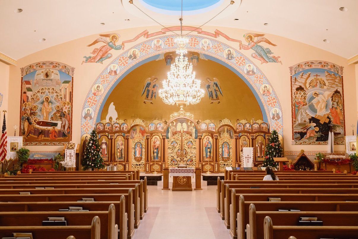 Ornate church interior with chandelier, painted arch, and rows of wooden pews facing the altar