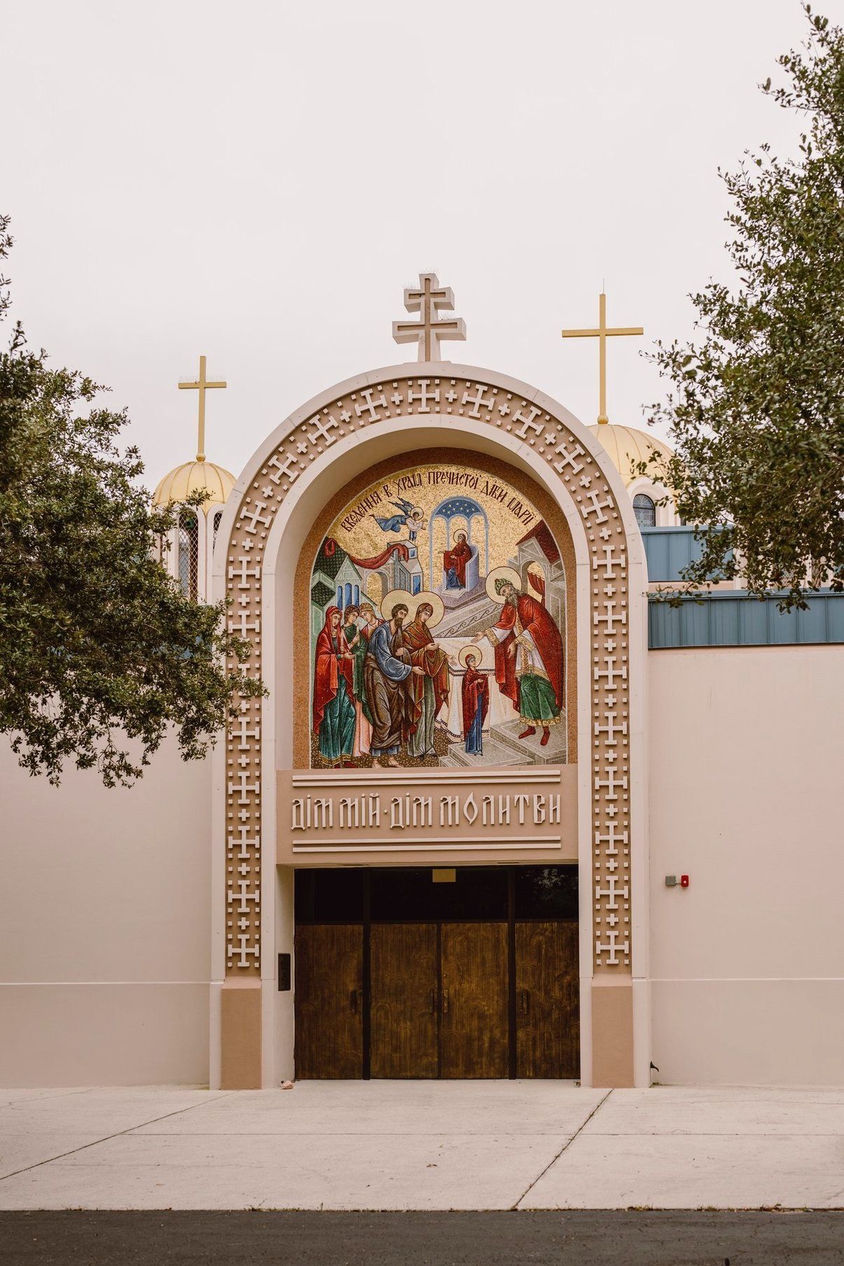 Church entrance with a colorful mosaic above wooden doors and two gold crosses on the roof.
