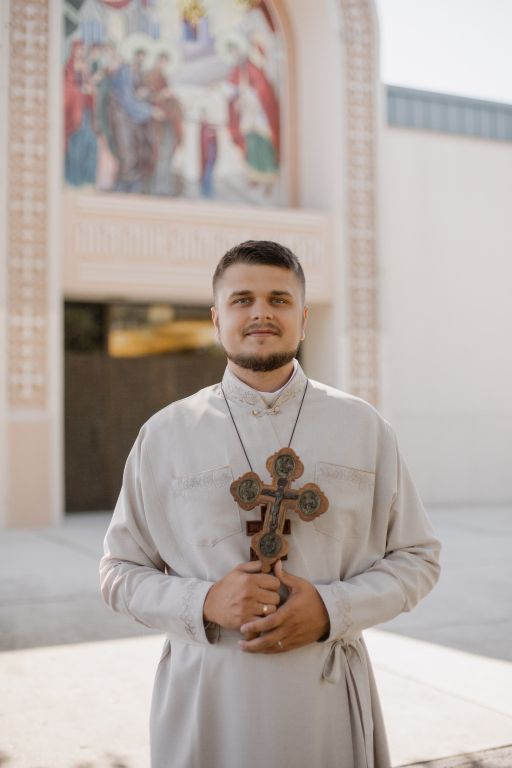 Cleric in white robes holds a cross in a church interior, standing before religious artwork.