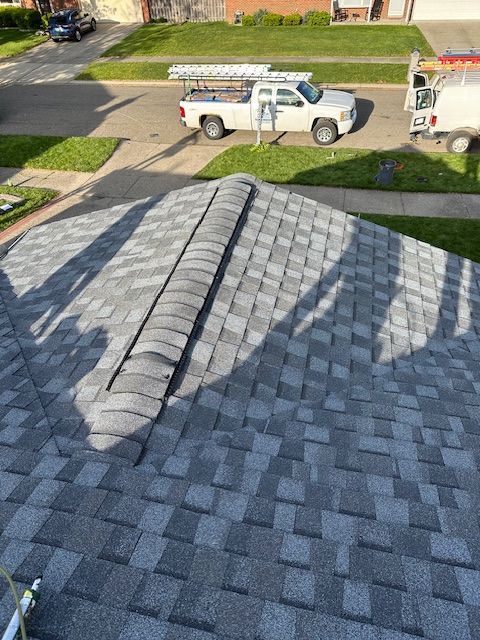 A view from a residential roof looking down at a gray shingled roof with a ridge cap, a white pickup truck, and a lawn. A view from a residential roof looking down at a gray shingled roof with a ridge cap, a white pickup truck, and a lawn.