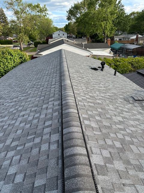 High-angle view of a grey shingled residential roof with a prominent ridge cap, looking down the peak toward houses. High-angle view of a grey shingled residential roof with a prominent ridge cap, looking down the peak toward houses.