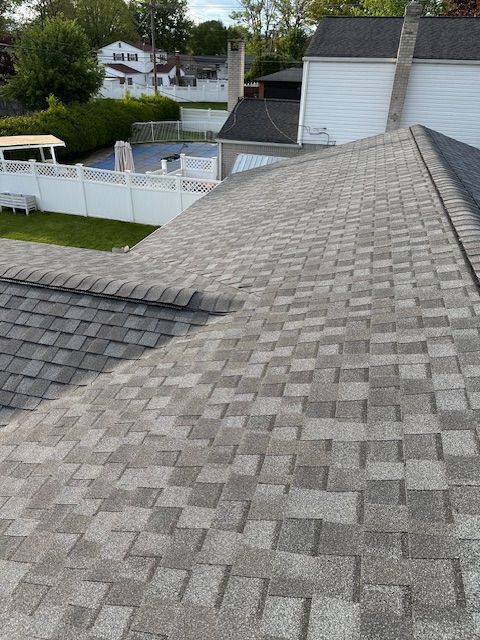 A high-angle view of a grey shingled roof, showing a intersecting roofline above a backyard with a white fence and lawn. A high-angle view of a grey shingled roof, showing a intersecting roofline above a backyard with a white fence and lawn.