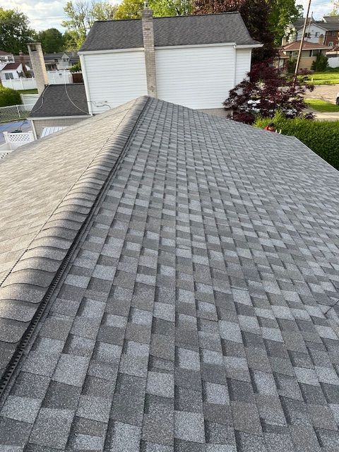 A view looking down over a gray asphalt shingle roof with a ridge cap, with a white garage building in the background. A view looking down over a gray asphalt shingle roof with a ridge cap, with a white garage building in the background.