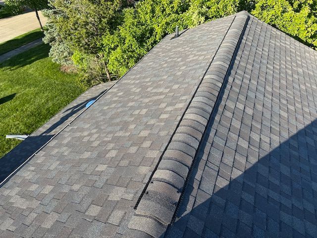 An elevated, high-angle view of a grey asphalt shingle roof with a ridge cap, bordered by green trees and a grassy lawn.