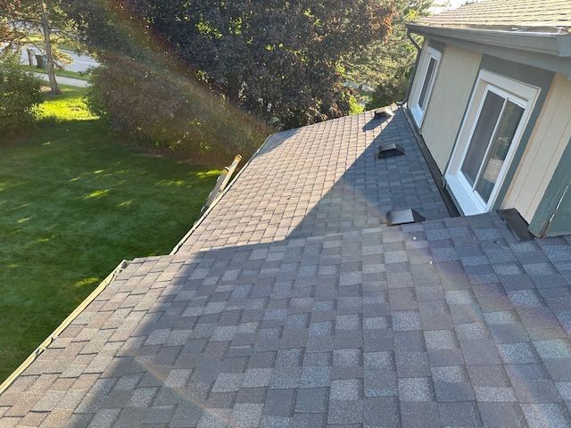 A high-angle view of a gray asphalt shingle roof on a house with two roof vents and a window visible on the side wall.