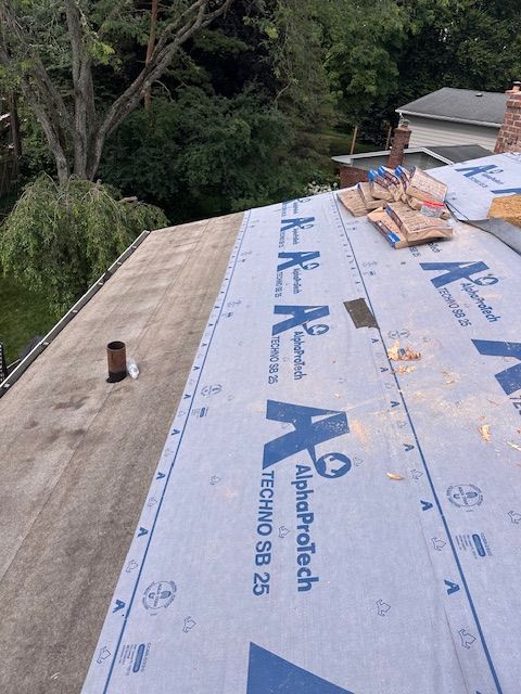 A residential roof under repair, featuring sections of exposed wood decking and blue synthetic underlayment.