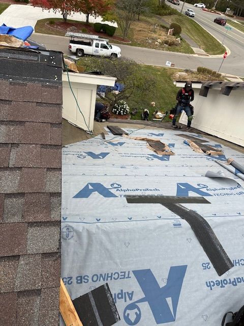 Roofers work on a residential roof covered with blue underlayment and shingles, seen from a high angle.