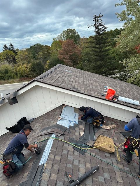 Two workers installing brown shingles on a residential roof against a cloudy sky background.