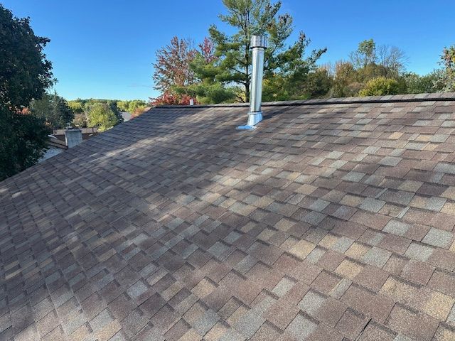 A high-angle view of a brown shingled roof with a single metal chimney vent under a clear blue sky.