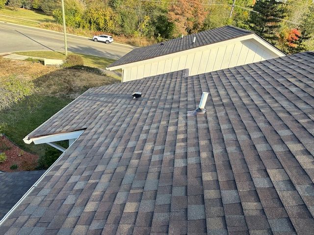 High-angle view of a brown shingled roof with two vent pipes, overlooking a neighborhood street and surrounding trees. High-angle view of a brown shingled roof with two vent pipes, overlooking a neighborhood street and surrounding trees.