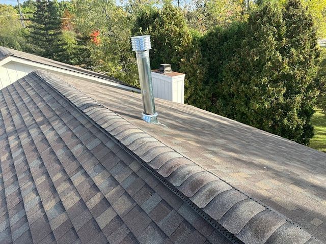 A metal vent pipe extending from a shingled roof, with a white chimney and green trees in the background.