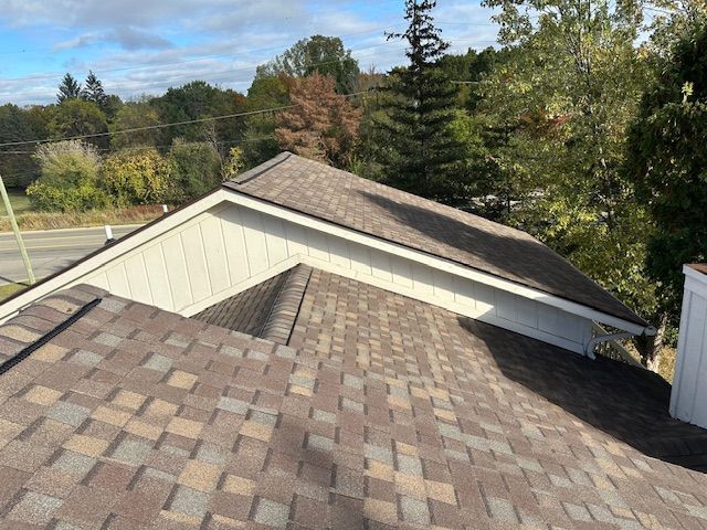 A high-angle view of a residential roof with brown asphalt shingles against a backdrop of trees and a blue sky.