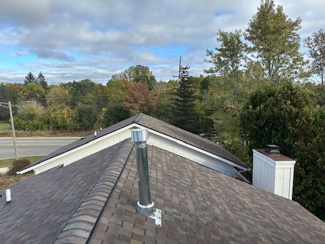 A residential roof with brown shingles, a metal chimney pipe, and a white chimney stack, set against trees and a cloudy sky.