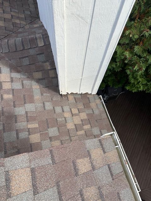 A close-up view of asphalt shingles on a roof meeting a white exterior wall corner above a gutter and dark decking.