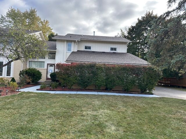 A two-story, cream-colored house with a dark shingled roof, a front lawn, and a large hedge row.