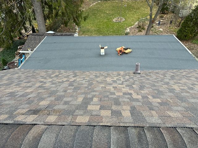 A view from a sloped shingled roof looking down onto a flat, grey roof section featuring a pipe and tools.