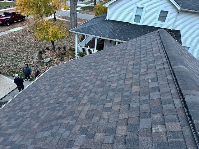 A high-angle view of a brown shingled roof, with three people standing on the ground near a suburban house.