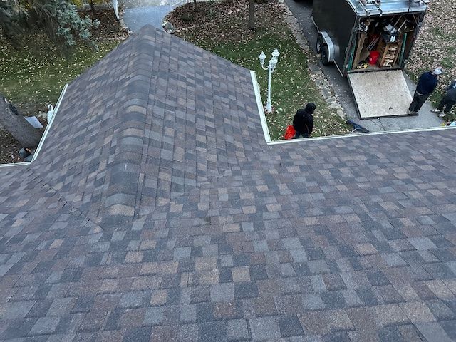 Workers on a roof during a construction or repair project, with a trailer parked on the driveway nearby.