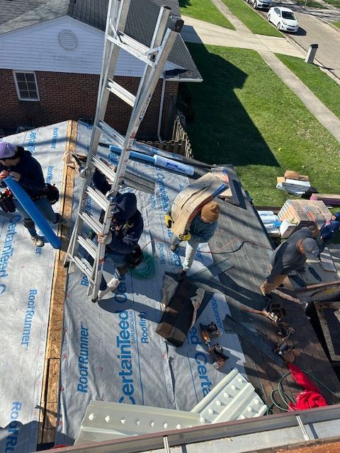 Roofers work on a residential roof covered with blue underlayment and shingles, seen from a high angle.