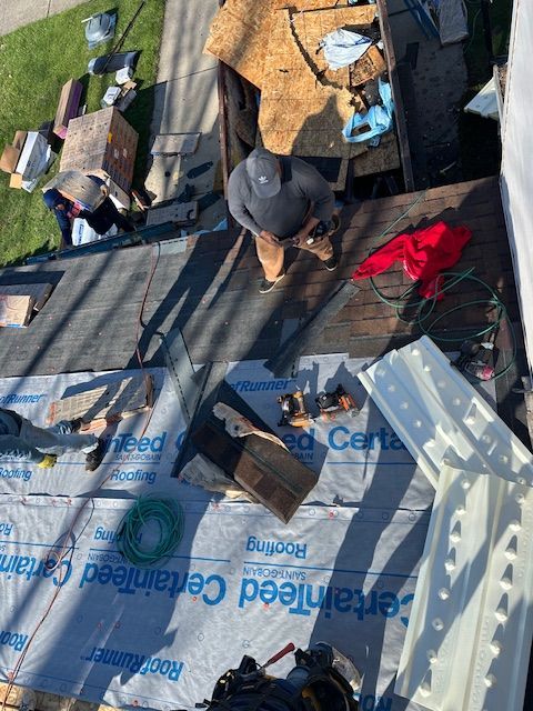 Two workers installing brown shingles on a residential roof against a cloudy sky background.