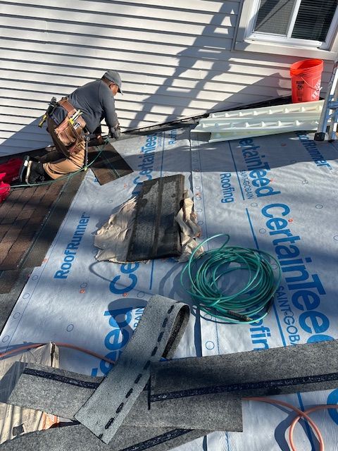 Worker installing roofing membrane on a house roof beside tools and coiled green hose
